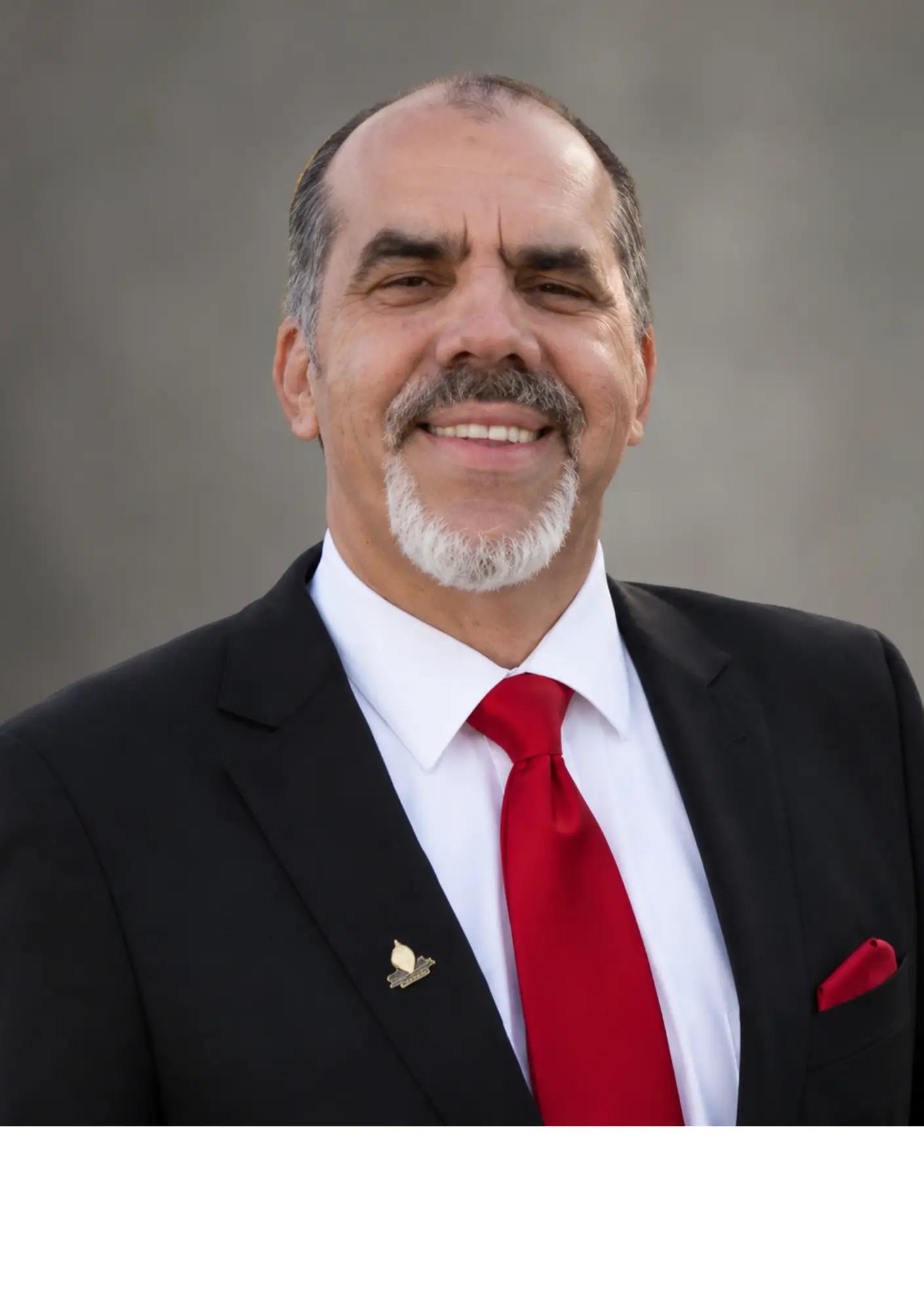 Smiling man in a black suit, white shirt, and red tie standing outdoors.